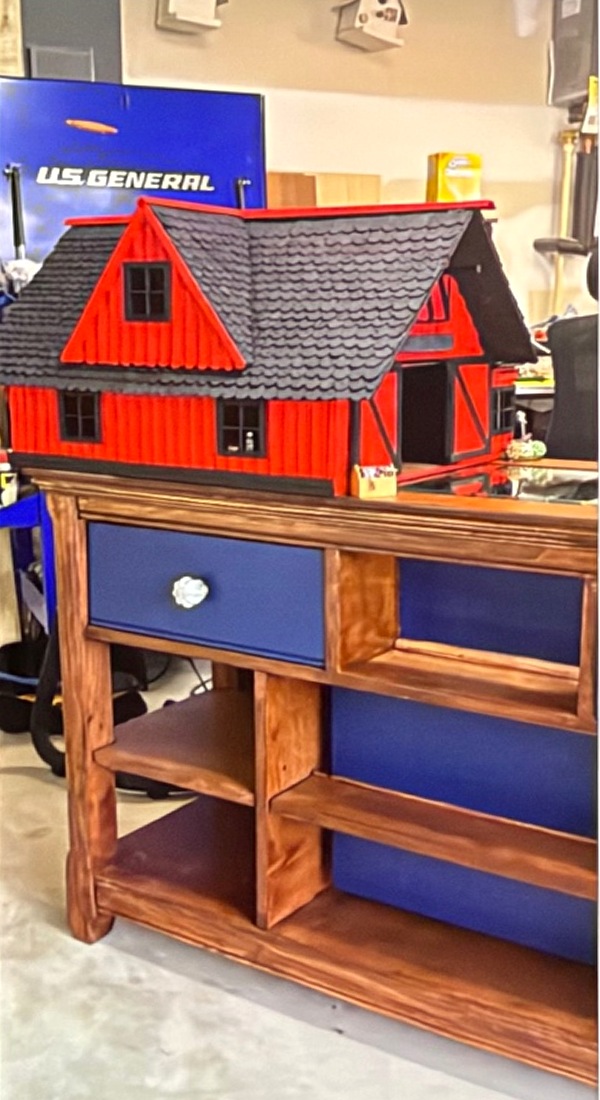 A red miniature barn with black trim and a textured shingle roof, sitting on an old dresser that hides one giant connected cat house inside.